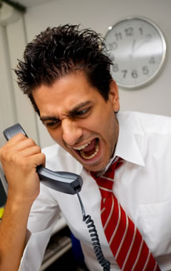 image of a visibly stressed male office worker sat at a desk yelling down a phone handset.